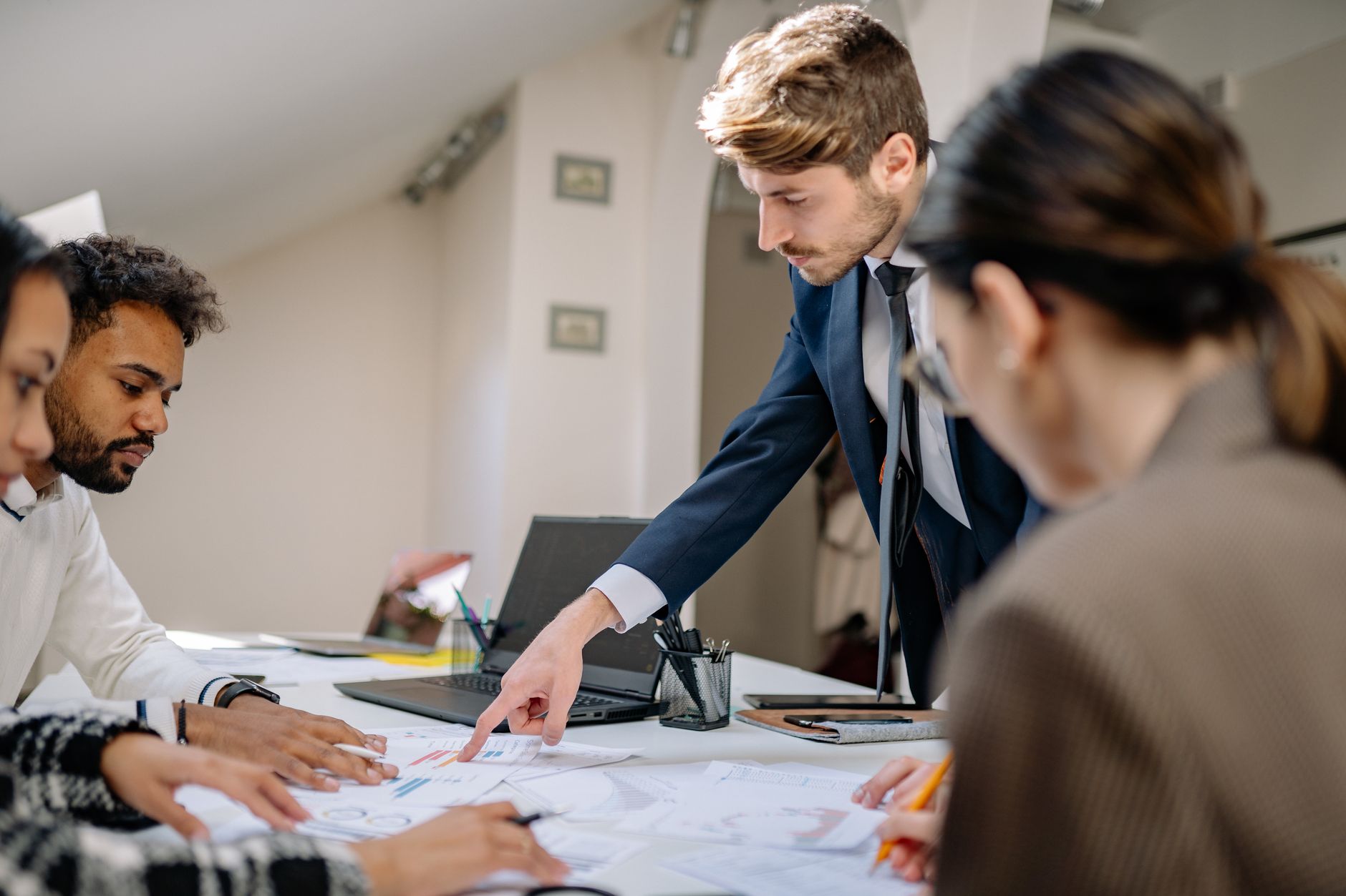 Équipe réunie autour d’une table de bureau : un homme en costume désigne des graphiques imprimés, près d’un ordinateur portable et d’un pot à stylos.