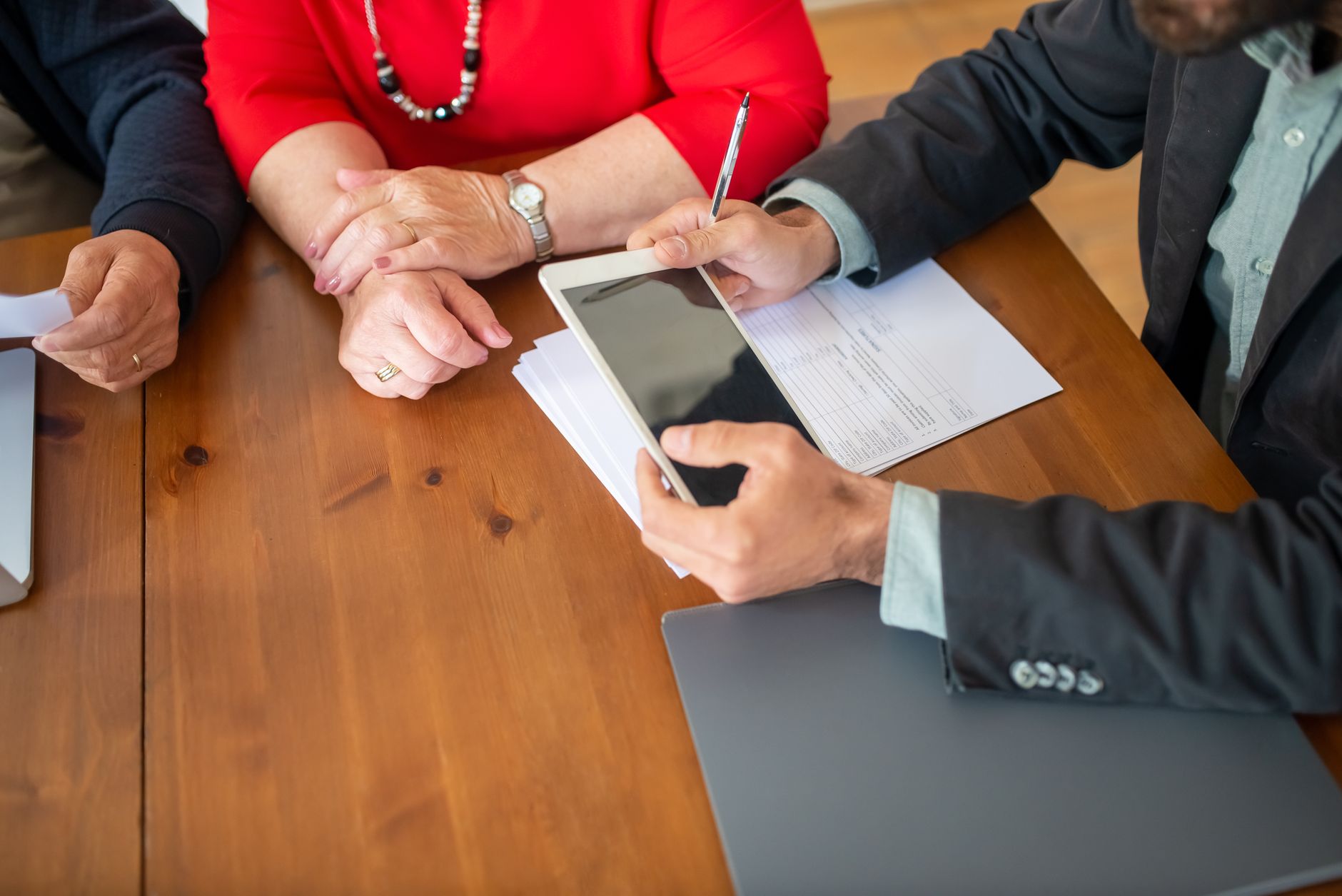 Rendez-vous autour d’une table en bois : un homme présente une tablette posée sur des documents à une femme en rouge, les mains jointes.