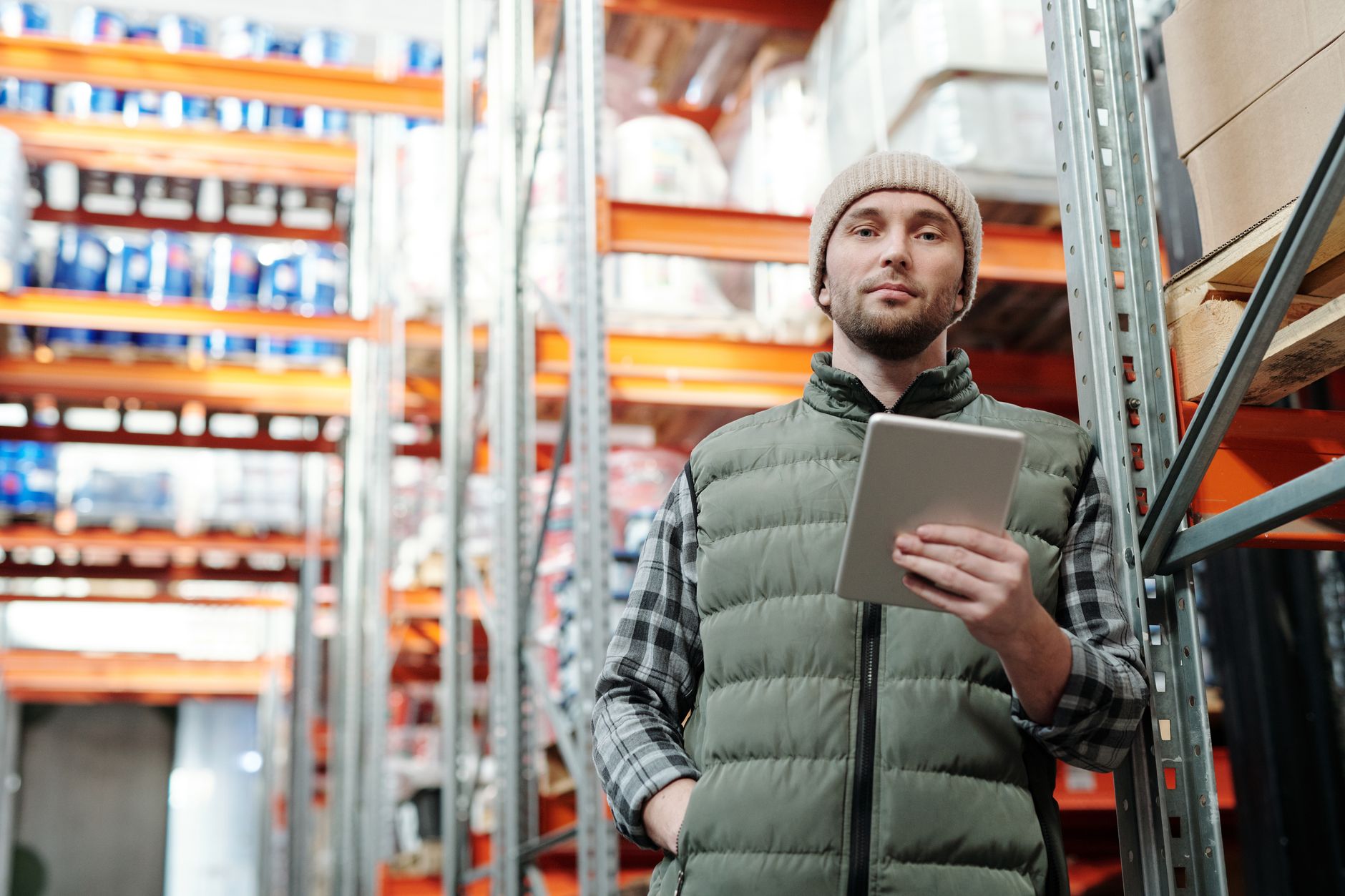 Un homme en bonnet et gilet vert tient une tablette dans un entrepôt, entre des rayonnages métalliques chargés de palettes et de cartons.