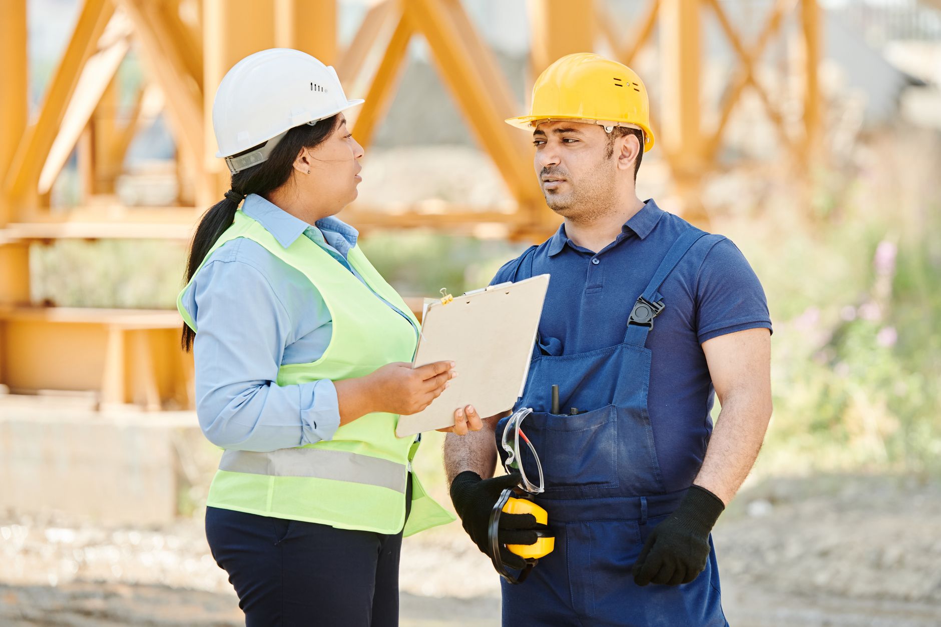 Une femme en casque blanc et gilet jaune tient un porte-bloc et parle à un homme en casque jaune et salopette bleue, sur un chantier.